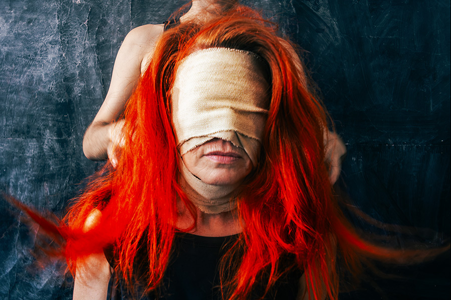 Nahaufnahme einer Frau mit langem rotem Haar und Augenbinde aus Stoff, Hände greifen von hinten ins Haar, dunkler strukturierter Hintergrund, konzeptuelle Fotografie zum Thema Imagination und Blindheit