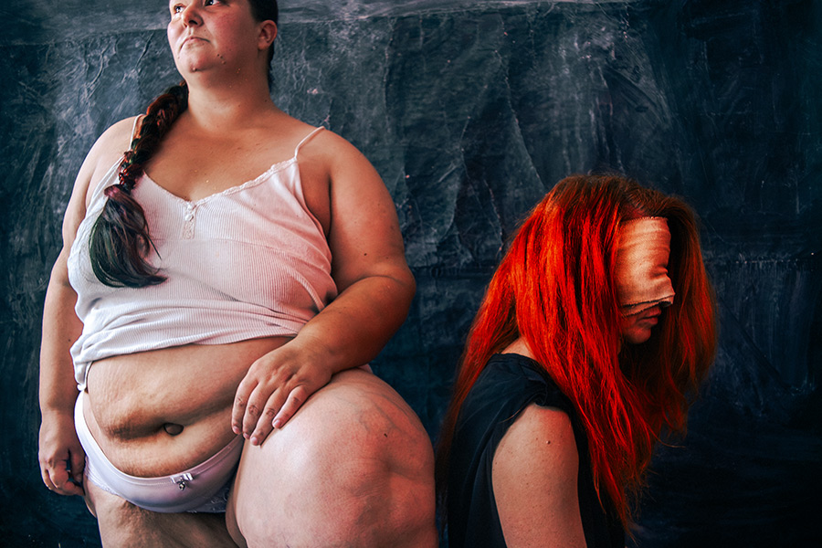 Zwei Frauen im Ganzkörperporträt nebeneinander, eine mit dickem, ungeschöntem Körper in weißer Unterwäsche, die andere mit rotem Haar und Augenbinde, Rücken an Rücken, minimalistischer dunkler Hintergrund, inszenierte Körperfotografie
