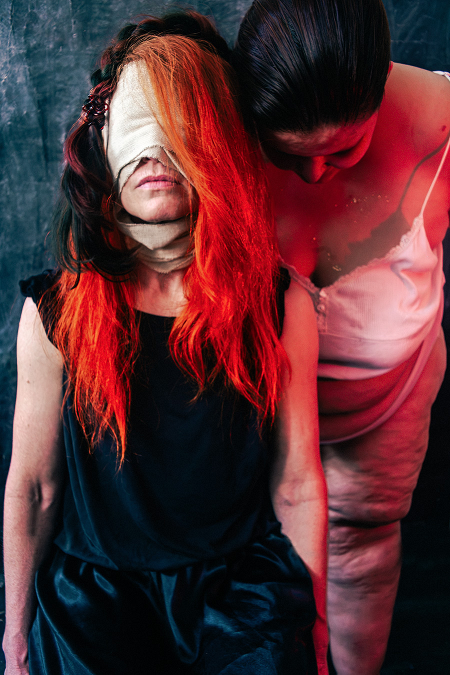 Zwei Frauen im Profil, verbundene Augen unter rotem Haar, zweite Figur mit dickem Körper und Träger-Unterhemd, dunkler Hintergrund, inszenierte Fine-Art-Fotografie, Thema Sehen und Nicht-Sehen