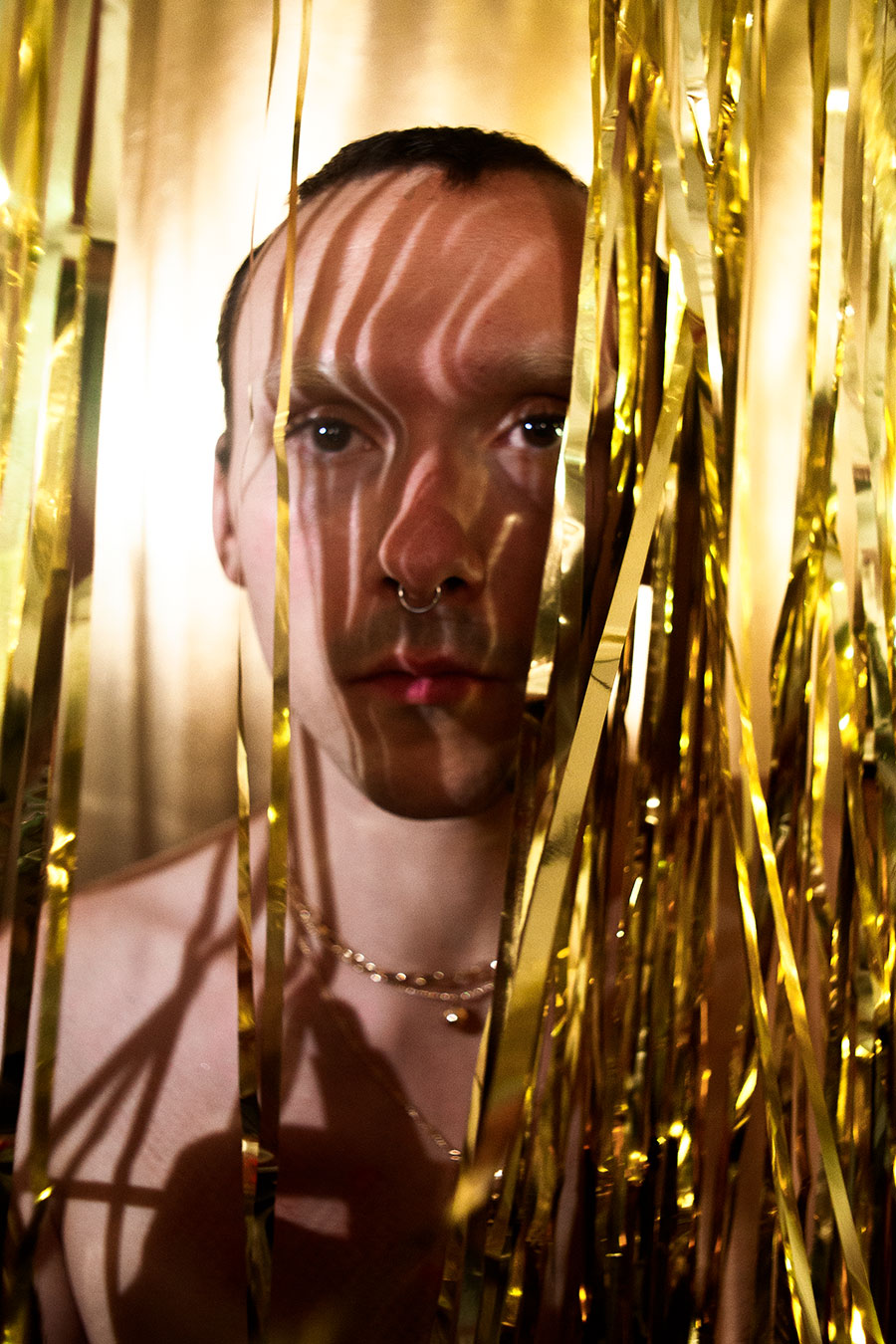 Halb verdecktes Gesicht hinter goldenen Lamettafäden, Streifenschatten auf der Stirn, Septum-Piercing, nackter Oberkörper, Kettenschmuck, analog-digitale Hybridaufnahme, konzeptuelle Porträtfotografie