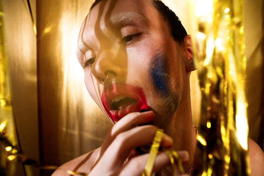 Profilaufnahme einer Person beim Auftragen von rotem Lippenstift, blaue Farbspuren an der Wange, Lamettavorhang im Hintergrund, inszenierter Schminkprozess als konzeptuelle Fotografie, analoges Pentacon-Objektiv