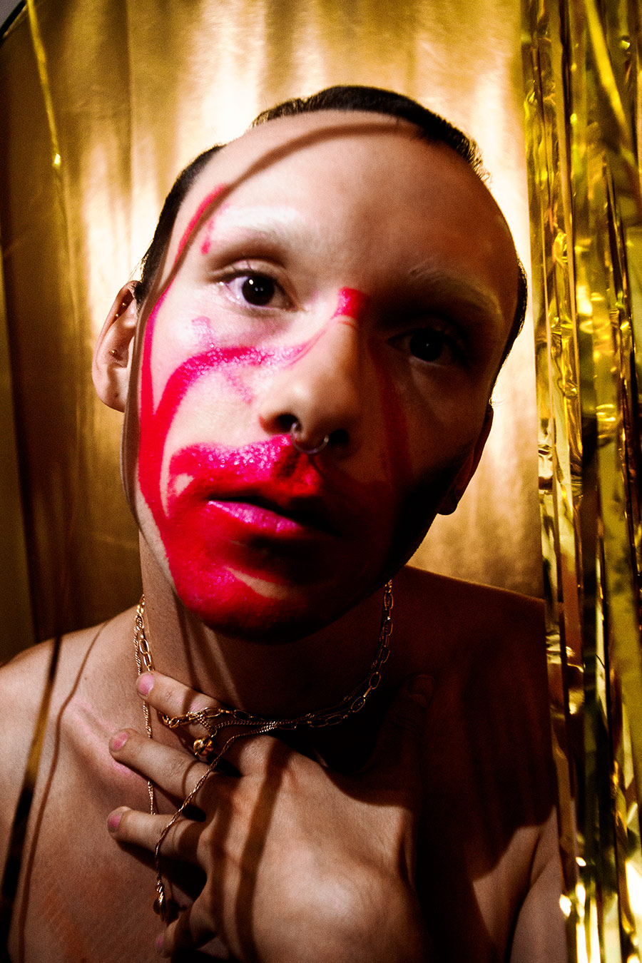 Extreme Nahaufnahme eines Gesichts mit roter, tränenartiger Bemalung, Septum-Piercing, Hand am Hals mit mehreren Goldketten, direkter Blick, goldener Lamettahintergrund, analoge Optik, Rausch-Serie