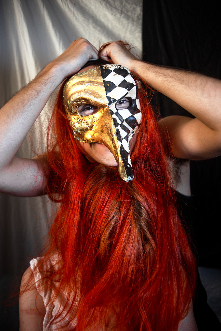 Frontalporträt mit venezianischer Gold-Schachbrett-Maske, langes rotes Haar, Hände greifen von hinten in die Haare, dunkler Hintergrund, analoge Pestarztmaske als Todesmetapher