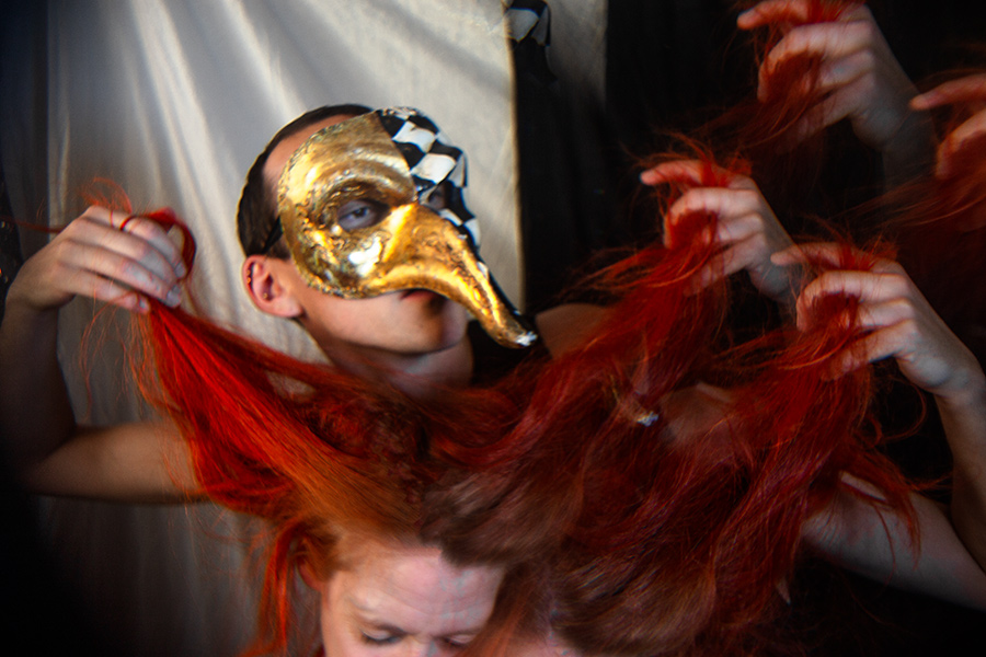 Person mit goldener Schachbrettmaske hält rote Haarsträhnen wie Fäden, mehrere Hände im Bild, heller Vorhang im Hintergrund, konzeptuelle Porträtfotografie Tod-Serie