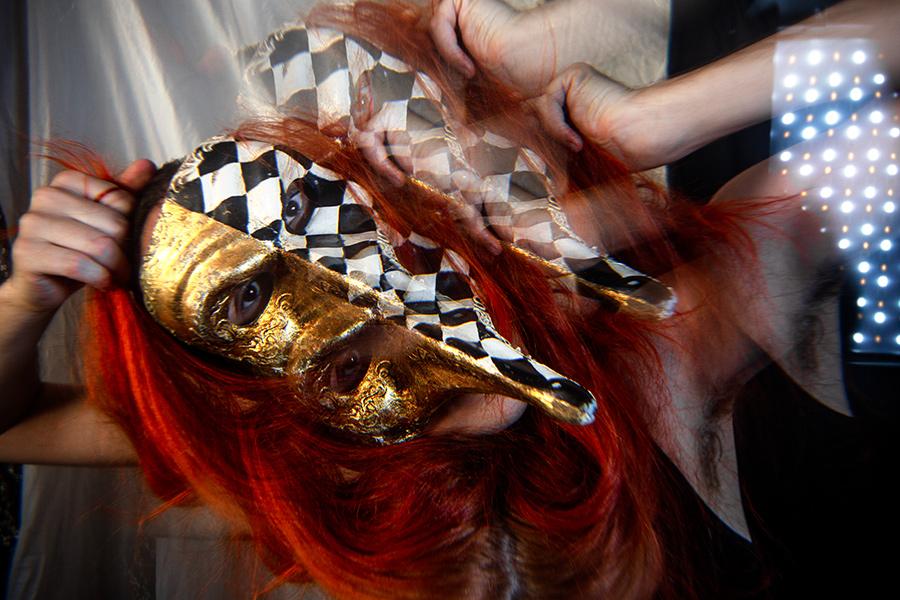 Verschwommene Doppelbelichtung einer goldenen Schnabelmaske, rotes Haar von Händen gehalten, Lichtpanel im Hintergrund, venezianische Maskerade als Todessymbol, inszenierte Fotokunst