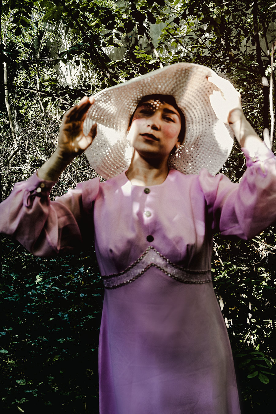 Frau hebt weißen Häkelhut mit beiden Händen über den Kopf, lila Vintage-Kleid, dunkler Waldhintergrund, inszenierte konzeptuelle Porträtfotografie