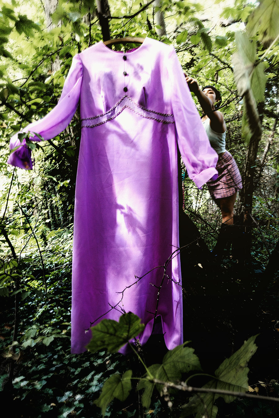 Lila Vintage-Kleid hängt auf Bügel an Baum im Wald, Frau klettert dahinter, Gegenlicht und Blattwerk, inszenierte konzeptuelle Farbfotografie