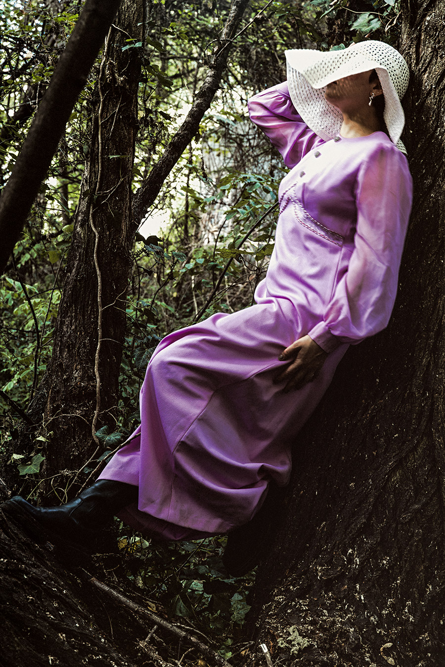 Inszenierte Porträtfotografie im Wald, Frau in fliederfarbenem Vintage-Kleid und weißem Häkelhut lehnt an Baumstamm, gepunktete Blattschatten, Farbfotografie, konzeptuelle Fotokunst „Der Garten der verzweigten Lichter"