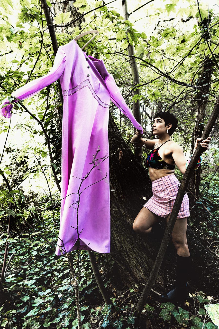 Inszenierte Fotografie mit an Baum hängendem lila Vintage-Kleid auf Bügel, Frau in Bikinitop und kariertem Rock klettert daneben, Wald, Farbfotografie