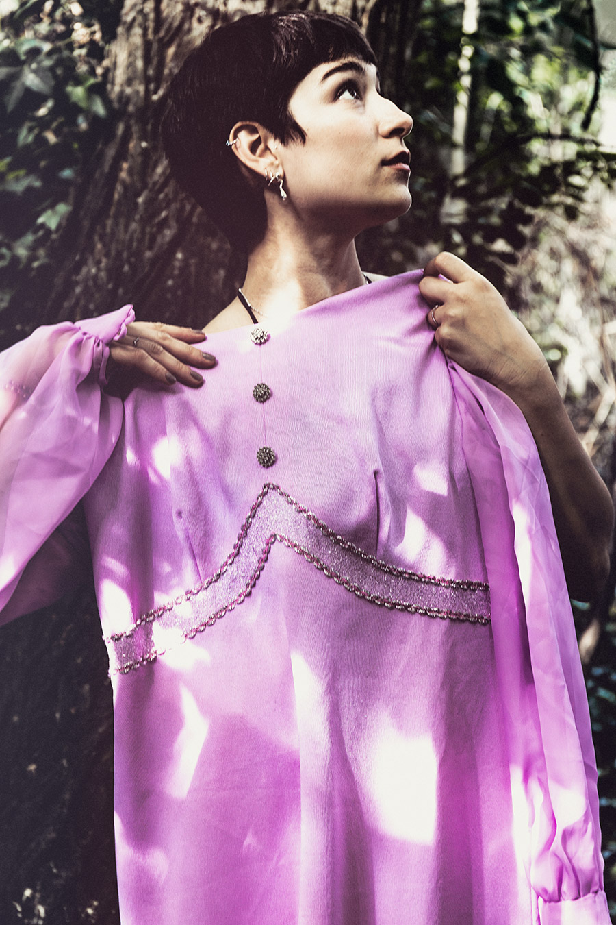 Waldporträt mit Sonnenflecken auf pinkfarbenem Vintage-Kleid, Frau hält Kleid an den Schultern, Blick nach oben, Perlenstickerei, inszenierte Farbfotografie