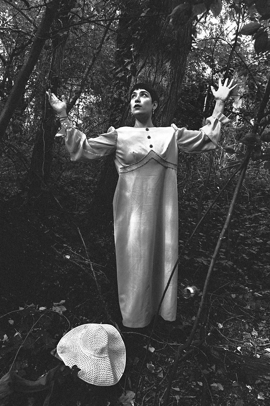 Schwarz-Weiß-Ganzkörperporträt einer argentinischen Tänzerin mit erhobenen Armen im Wald, Häkelhut am Boden, Vintage-Kleid, dramatisches Licht, konzeptuelle Fotoserie