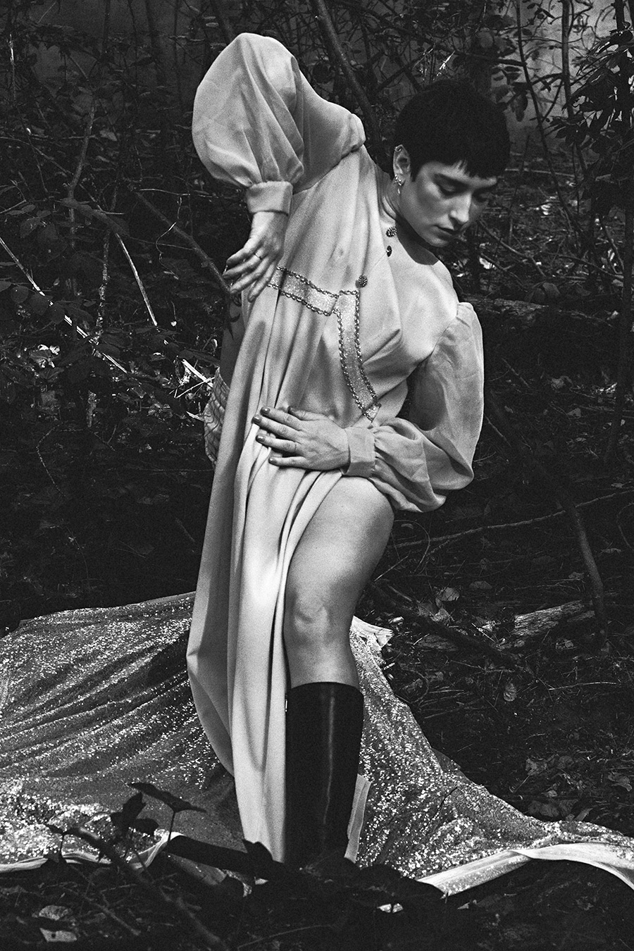 Schwarz-Weiß-Porträt einer argentinischen Tänzerin im Wald, Kleid über den Kopf gezogen, schwarze Stiefel, silberne Pailletten-Decke am Boden, konzeptuelle Fotografie