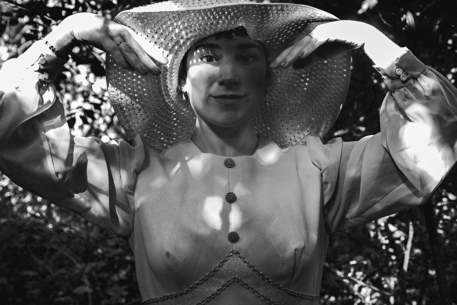 Schwarz-Weiß-Porträt einer argentinischen Tänzerin im Wald, hält weißen Häkelhut mit beiden Händen über den Kopf, Vintage-Kleid mit Perlenstickerei, Lichtflecken auf der Haut, konzeptuelle Fotoserie „Der Garten der verzweigten Lichter"