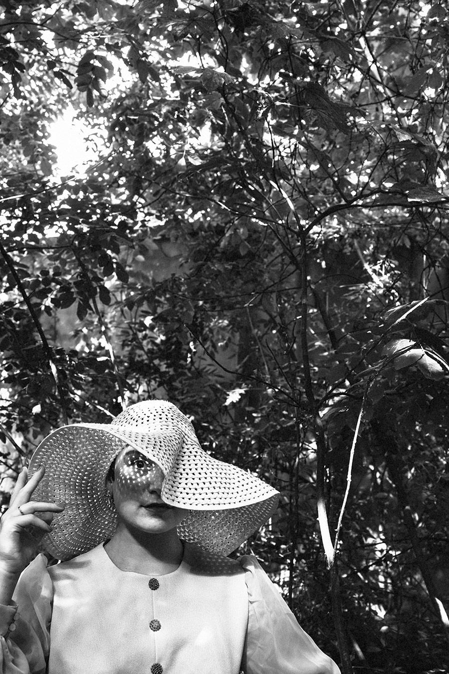 Schwarz-Weiß-Porträt einer argentinischen Tänzerin im Wald, ein Auge durch Löcher im Häkelhut sichtbar, Vintage-Kleid, dichtes Blätterdach im Hintergrund, konzeptuelle Fotografie