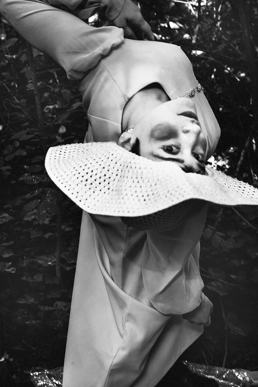 Schwarz-Weiß-Porträt einer argentinischen Tänzerin kopfüber im Wald, Häkelhut verrutscht über das Gesicht, Vintage-Kleid, Bewegungsunschärfe, konzeptuelle Fotoserie