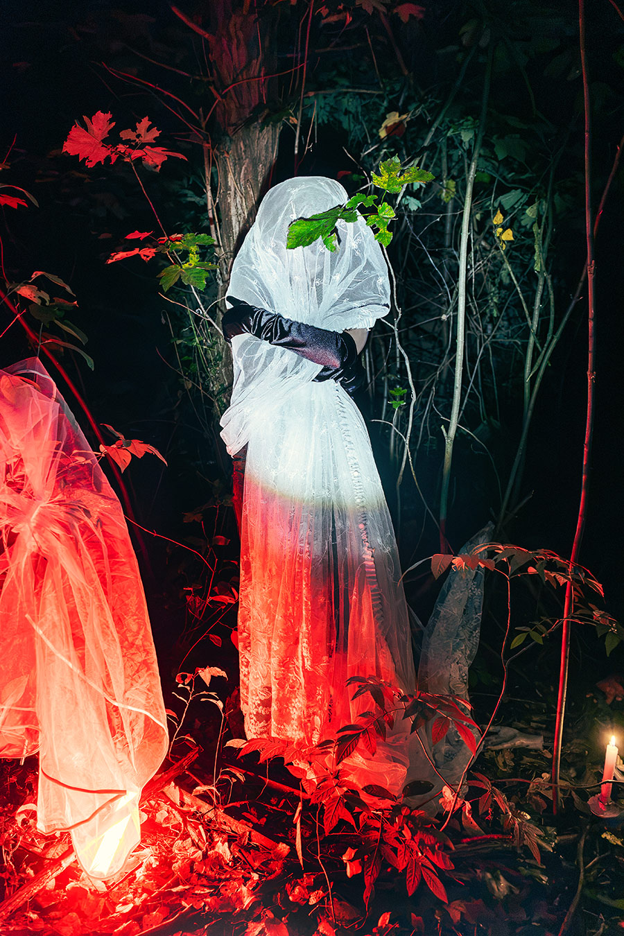 verhüllte Figur in weißer Spitze, Arme verschränkt, Blatt vor dem Gesicht, unten rotes Licht, Kerze im Unterholz, inszenierte Waldfotografie in der Dämmerung