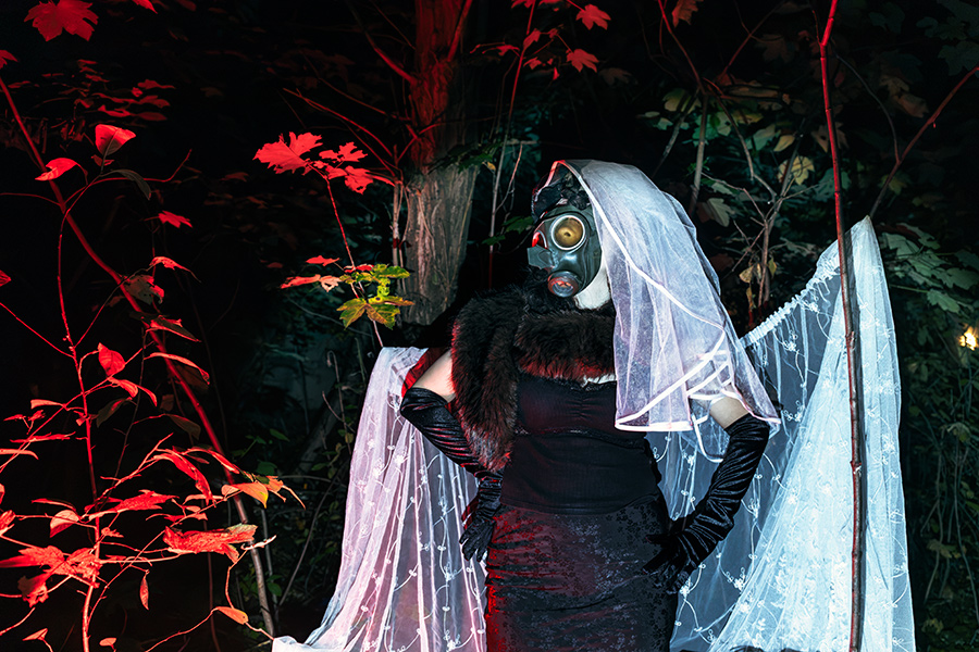 inszeniertes Porträt in der Dämmerung, Figur mit Gasmaske unter weißem Brautschleier, Pelzstola und schwarze Handschuhe, Spitzenvorhang im Wald, rotes Herbstlaub, buntes Kunstlicht, Serie Потёмки