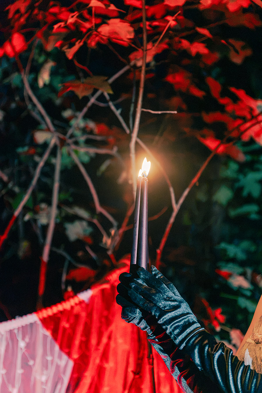 Detailaufnahme, schwarz behandschuhte Hand hält zwei schwarze Stabkerzen mit Flamme, rotes Herbstlaub, buntes Kunstlicht, inszenierte Dämmerung