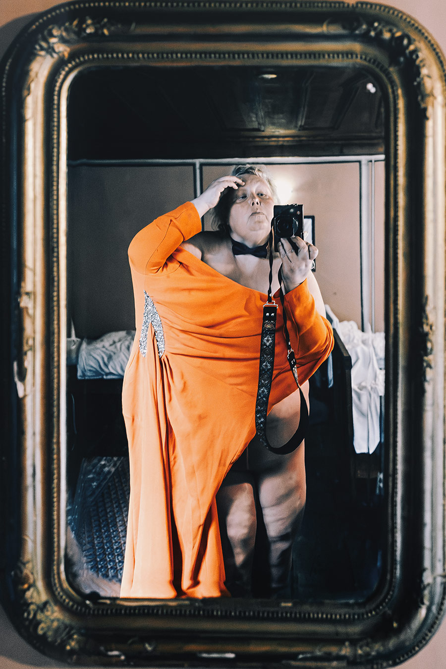 inszeniertes Selbstporträt im vergoldeten Spiegel, dicker Körper im orangenen Kleid mit Strassapplikation und Fliege, Hand im Haar, Leica in der Hand, Serie октомври
