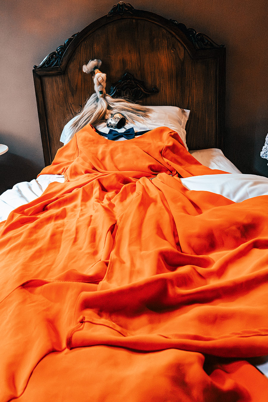 inszeniertes Stillleben, orangenes Kleid wie ein liegender Körper im Holzbett ausgebreitet, blauer Fliege und Pferdeschwanz mit Glocke auf dem Kissen, Fotokunst Leica, Serie октомври